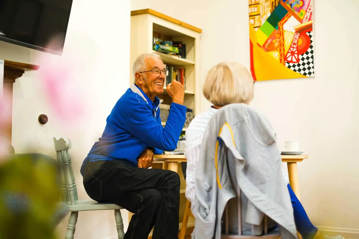 A man sitting at a table in a cafe with a book shelf and a picture of modern art with a lady whose back is to us. He has a blue jumper and is laughing and chatting.