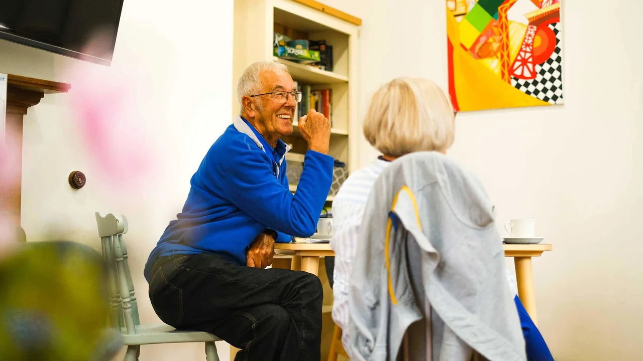 A man sitting at a table in a cafe with a book shelf and a picture of modern art with a lady whose back is to us. He has a blue jumper and is laughing and chatting.