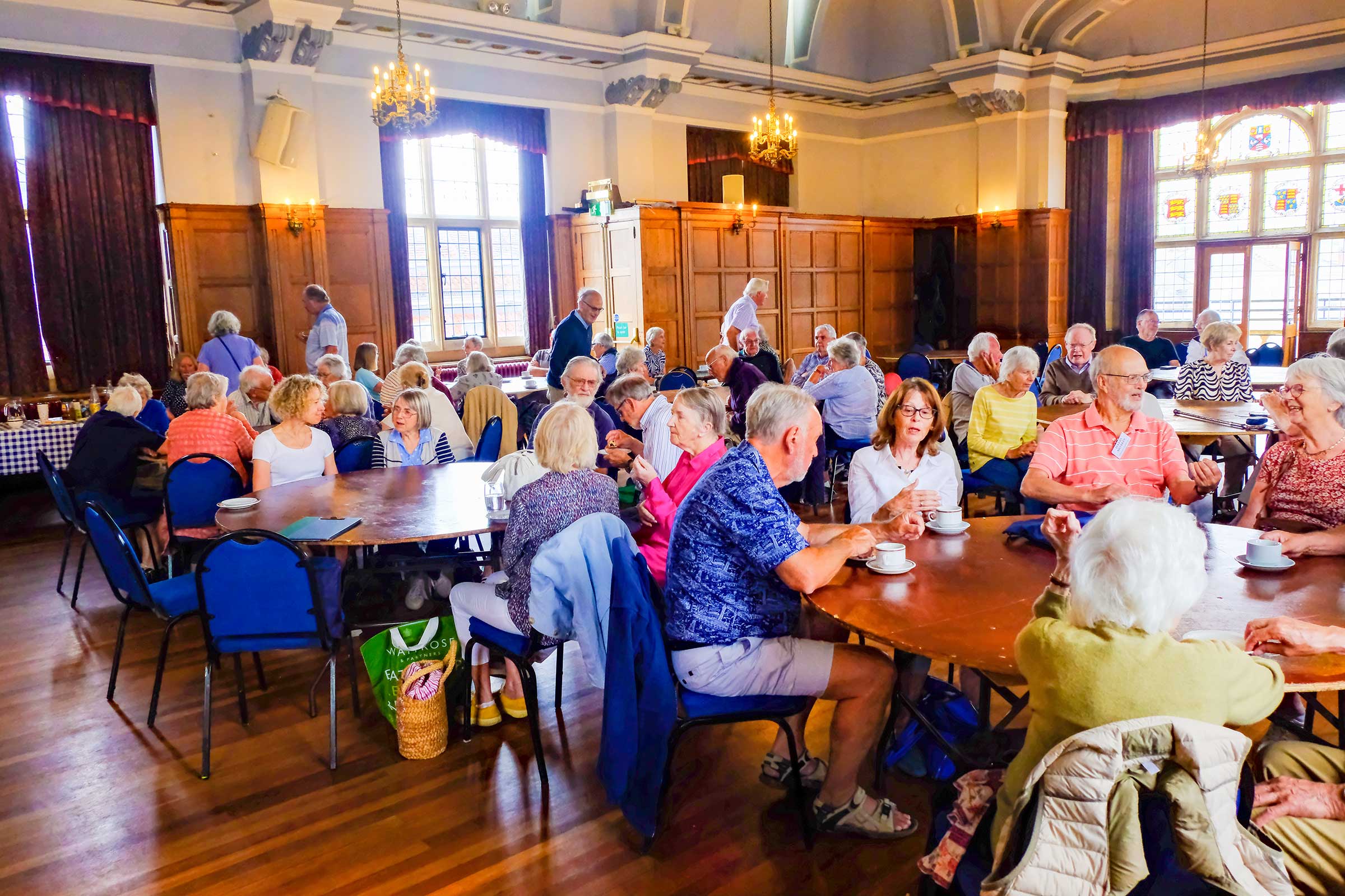 The picture shows a large room with grand high ceilings, a wooden floor and large stained glass windows, where many people are seated around round tables chatting and drinking tea or coffee