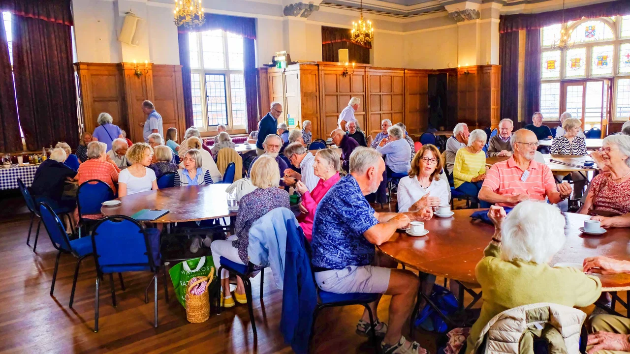 The picture shows a large room with grand high ceilings, a wooden floor and large stained glass windows, where many people are seated around round tables chatting and drinking tea or coffee