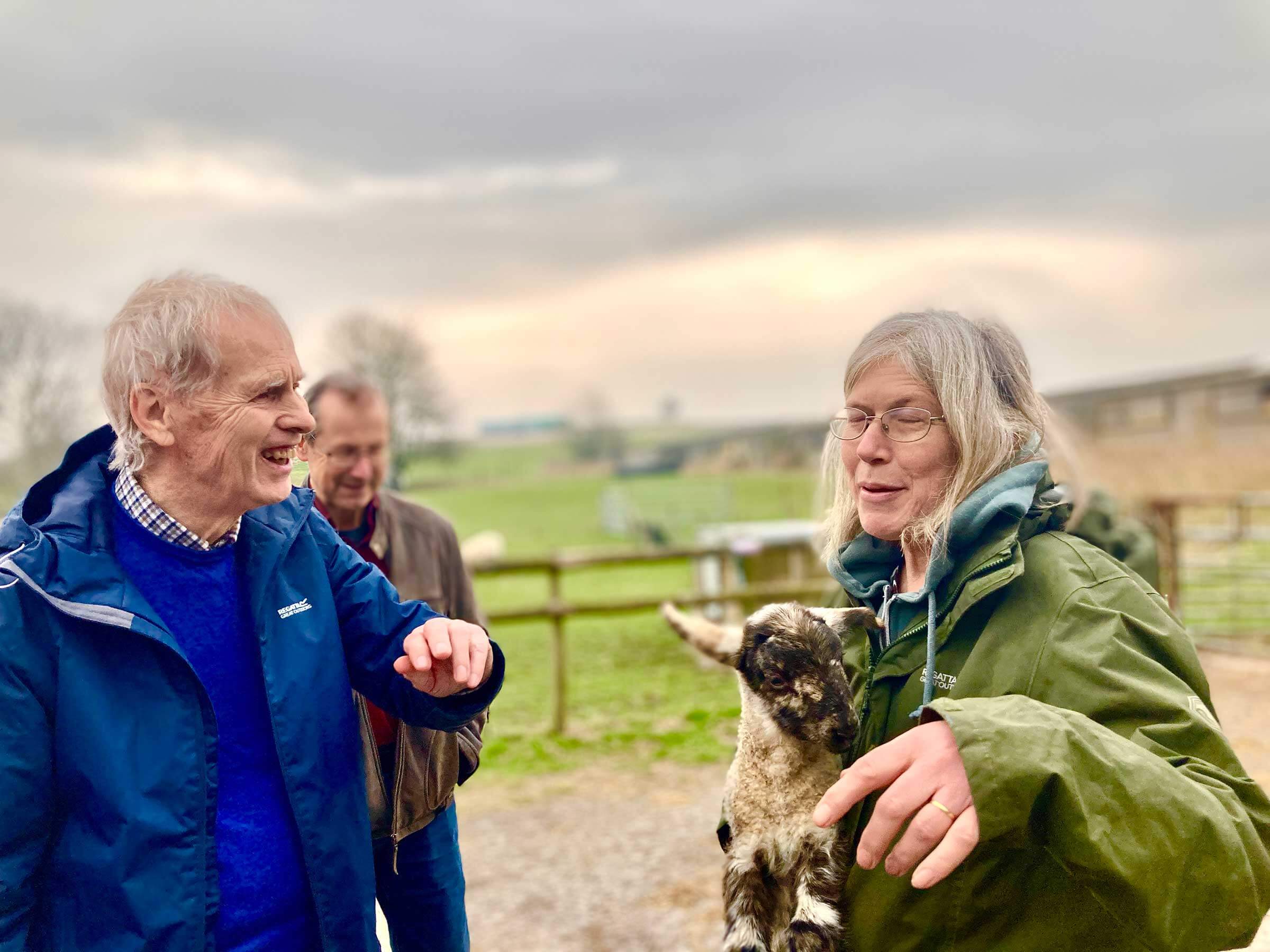 A older man wearing a blue anorak standing laughing with a lady in a green coat who is holding a lamb in her arms.  Taken at Kennet Muddy Boots group 