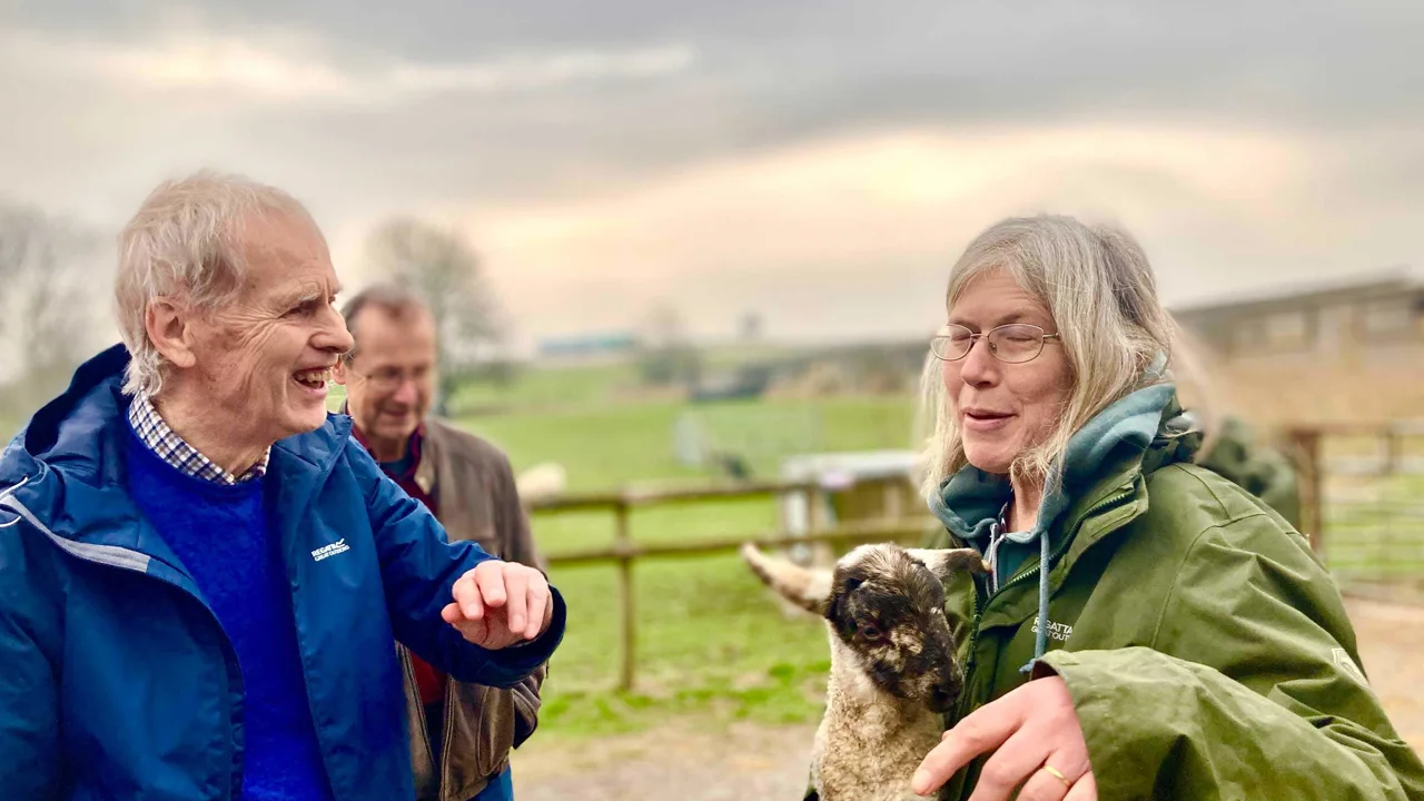 A older man wearing a blue anorak standing laughing with a lady in a green coat who is holding a lamb in her arms. Taken at Kennet Muddy Boots group