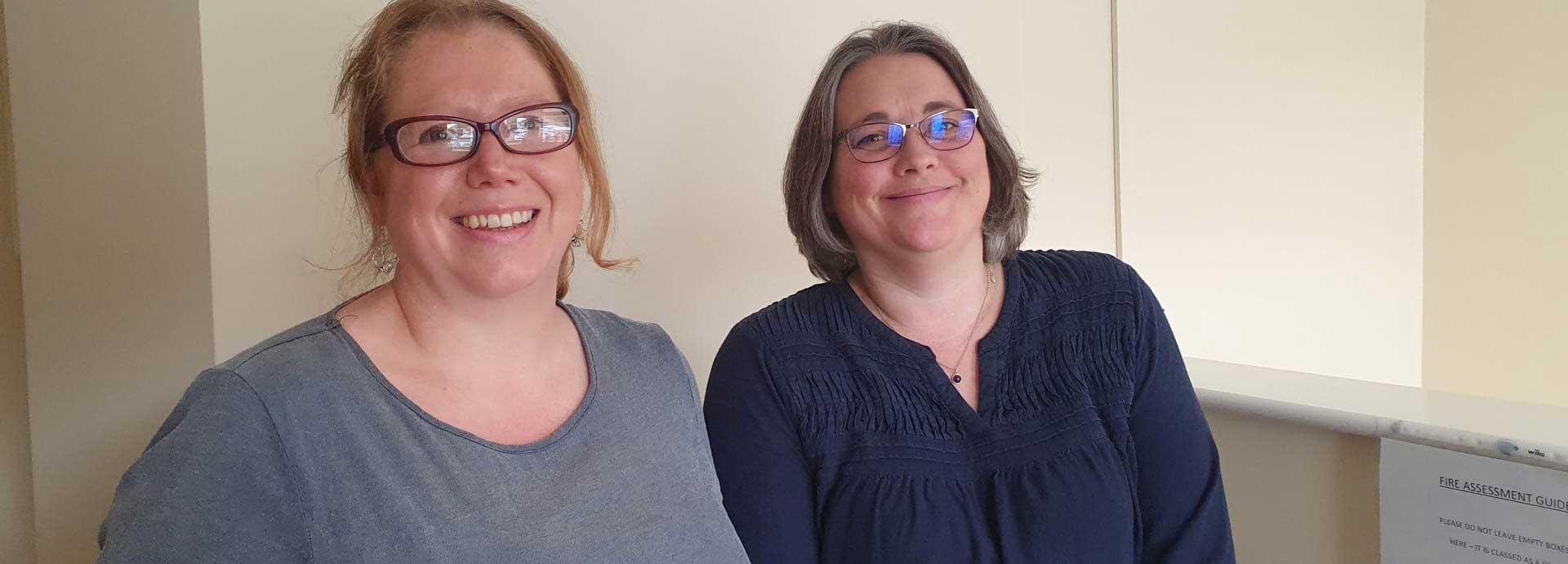 Close up of Sarah and Amy both wearing blue tops and smiling in an office 
