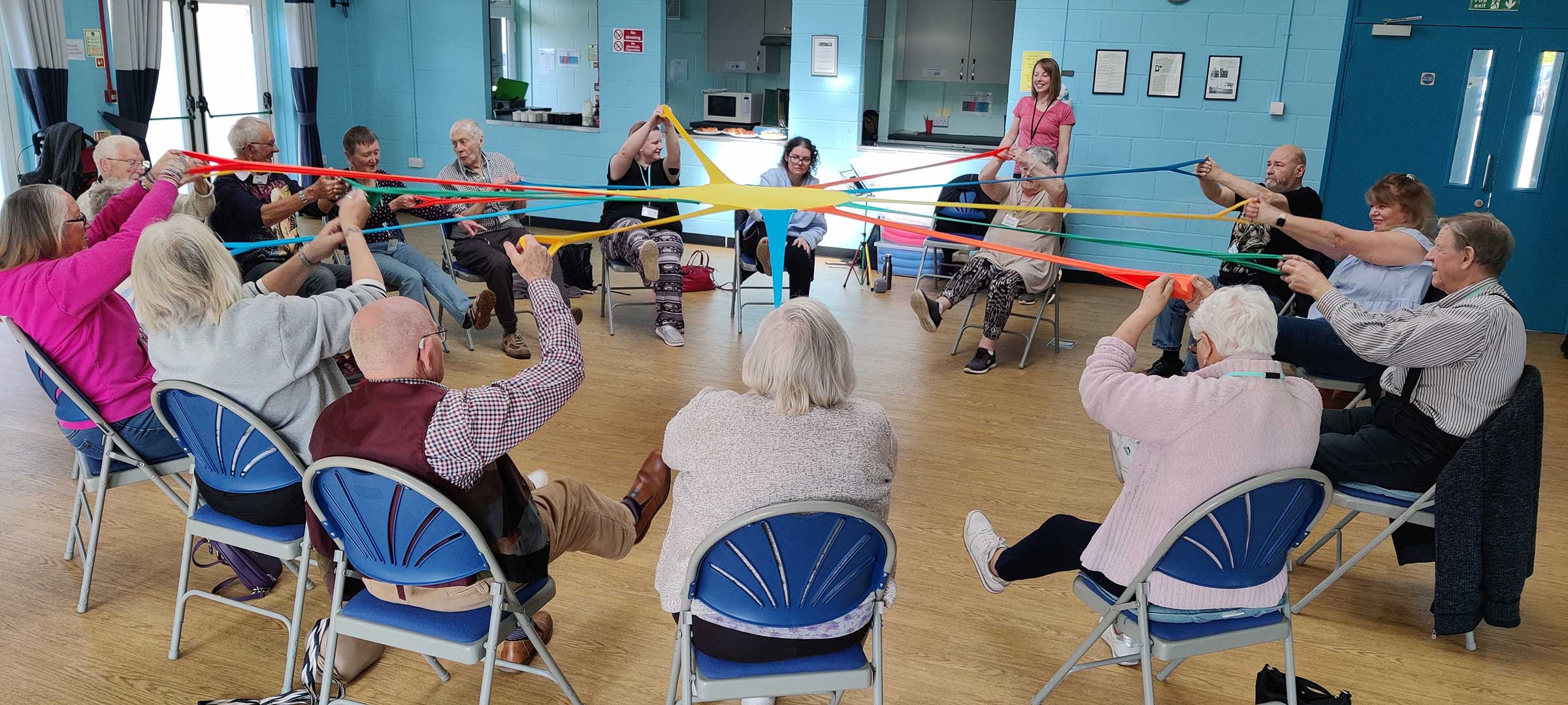 Sixteen older people sitting in a group pulling on red, yellow, green and blue stretchy bands joined in the middle all smiling at Melksham Movement for the Mind