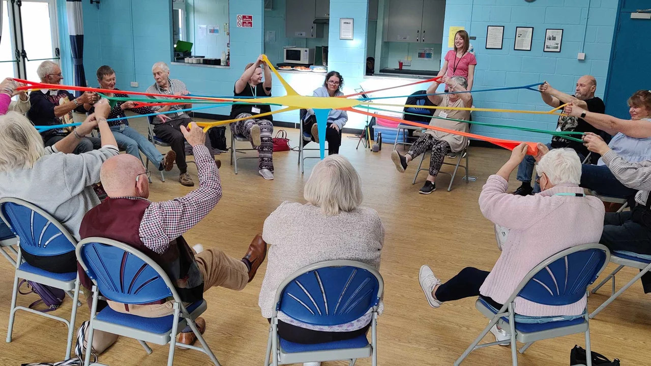 Sixteen older people sitting in a group pulling on red, yellow, green and blue stretchy bands joined in the middle all smiling at Melksham Movement for the Mind