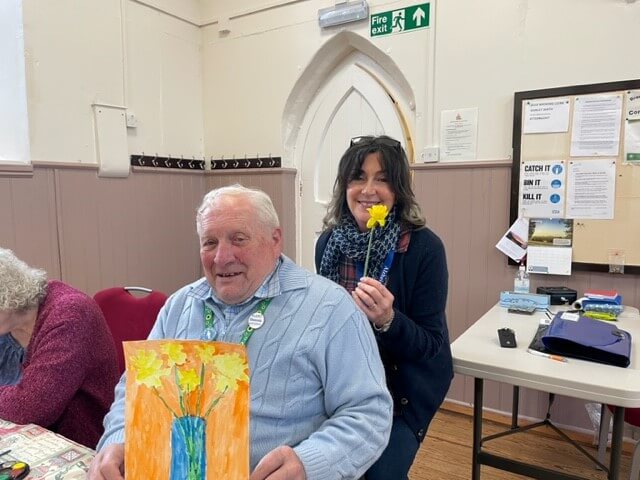 A gentleman in a blue jumper holds up a painting with orange background, a blue vase with five yellow daffodils.  Behind him a lady in black top holds up a yellow daffodil and is smiling.  Taken at Lyneham Art Group 