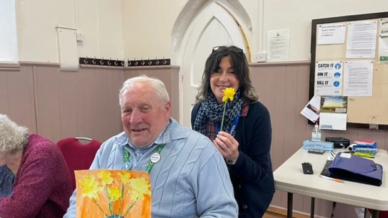 A gentleman in a blue jumper holds up a painting with orange background, a blue vase with five yellow daffodils. Behind him a lady in black top holds up a yellow daffodil and is smiling. Taken at Lyneham Art Group