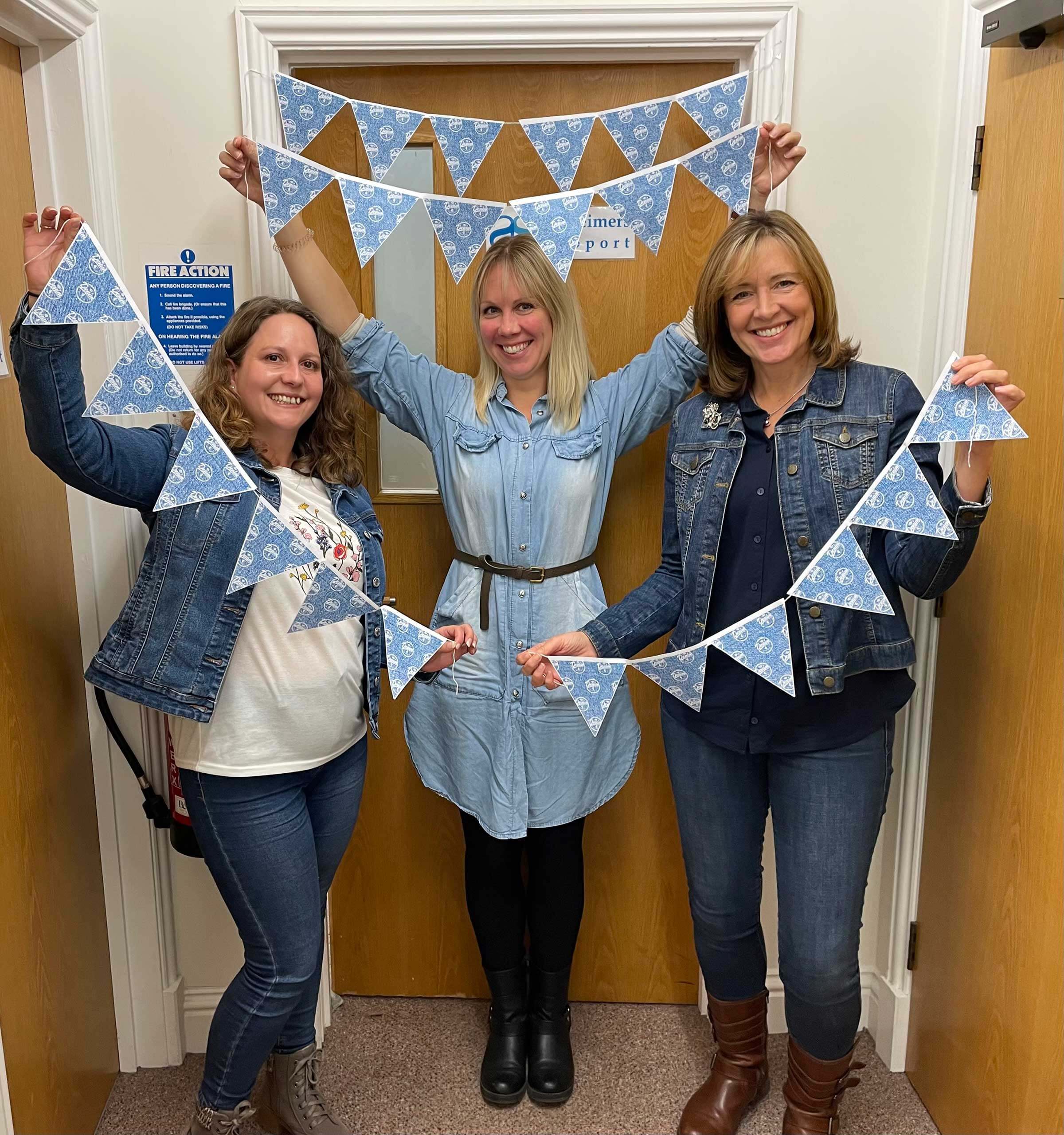 Denim 4 Dementia ladies in the office all wearing denim, smiling and holding up bunting 