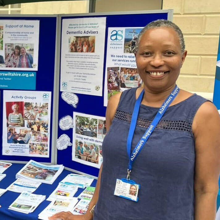 Annie Clayton in a blue sleeveless top, standing in front of an info board and next to a table carrying charity leaflets