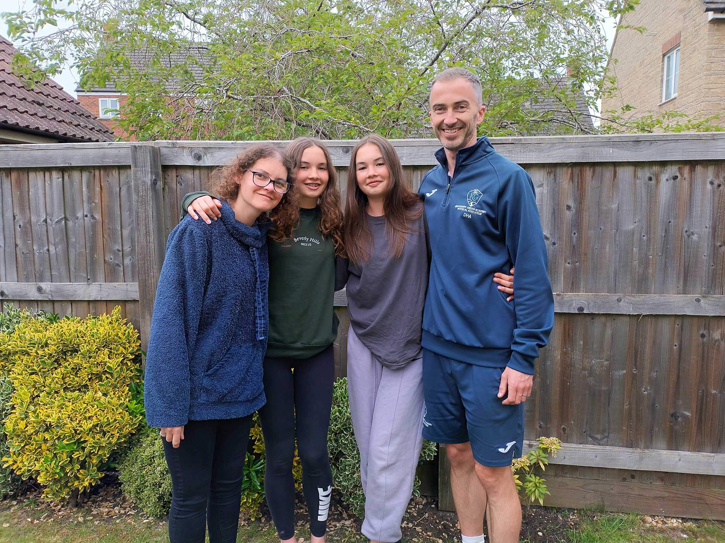 Claire and her family standing in a group with their arms around each other, in a garden setting in front of a wooden fence